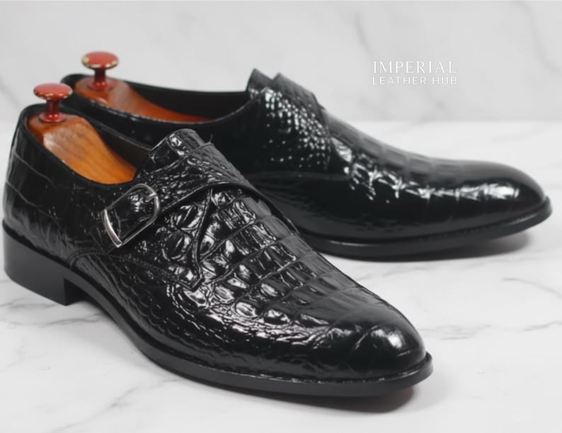 May include: A pair of black leather dress shoes with a crocodile skin texture. The shoes have a buckle closure and are displayed with wooden shoe trees. The shoes are on a white surface.