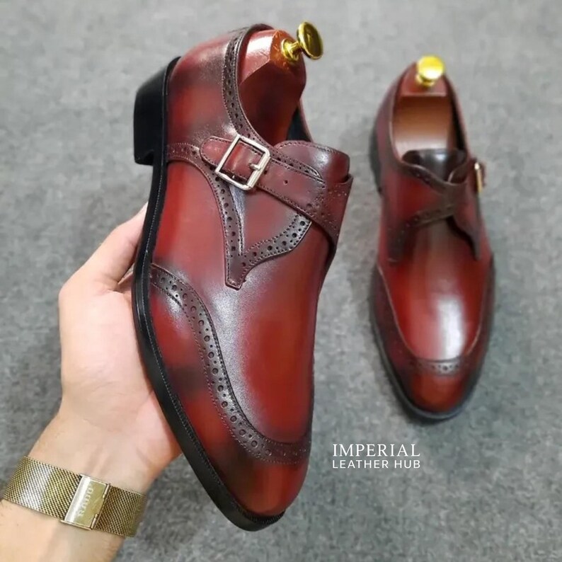 May include: A pair of deep red leather monk strap shoes featuring a burnished finish, brogue detailing along the seams, a silver-toned metal buckle, and a black sole. The text IMPERIAL LEATHER HUB is visible in the lower right corner of the image.