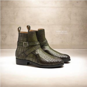 Op de afbeelding: Een paar olijfgroene enkellaarzen met een textuur van krokodillenleer, een zijband met een zilverkleurige metalen gesp en een treklipje bij de hiel. De tekst IMPERIAL LEATHER HUB is zichtbaar in de rechterbovenhoek van de afbeelding.