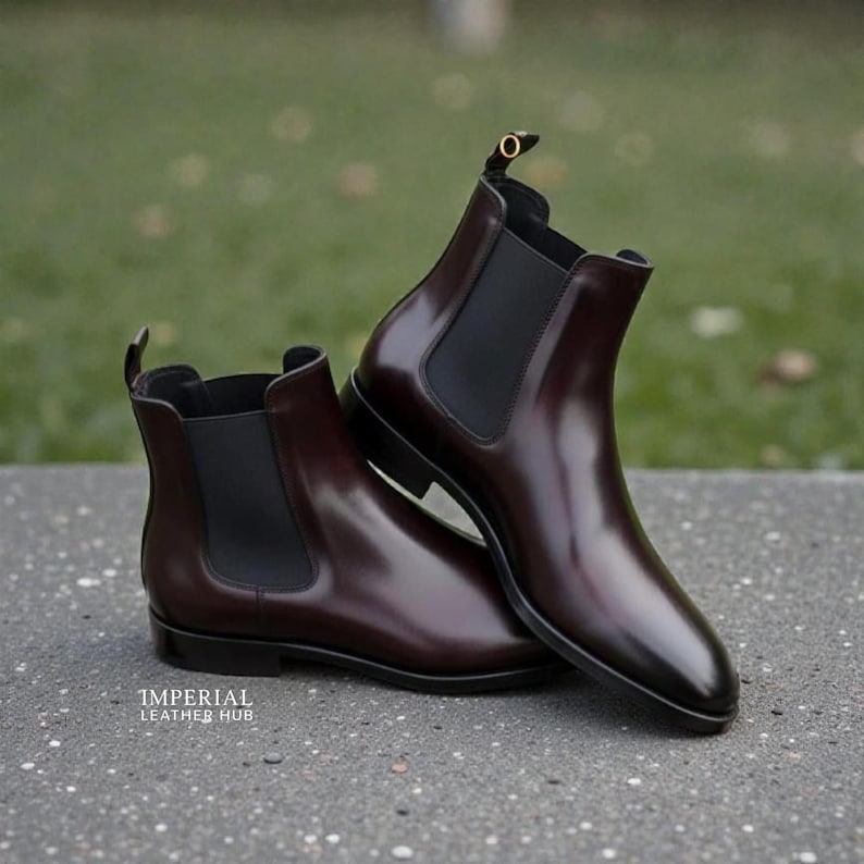 Pu&ograve; includere: Un paio di stivaletti Chelsea bordeaux con pannelli laterali elastici neri. Gli stivaletti hanno una finitura in pelle lucida e un tacco basso. Gli stivaletti sono esposti su una superficie grigia con uno sfondo verde sfocato. Il testo "IMPERIAL LEATHER HUB" &egrave; visibile.