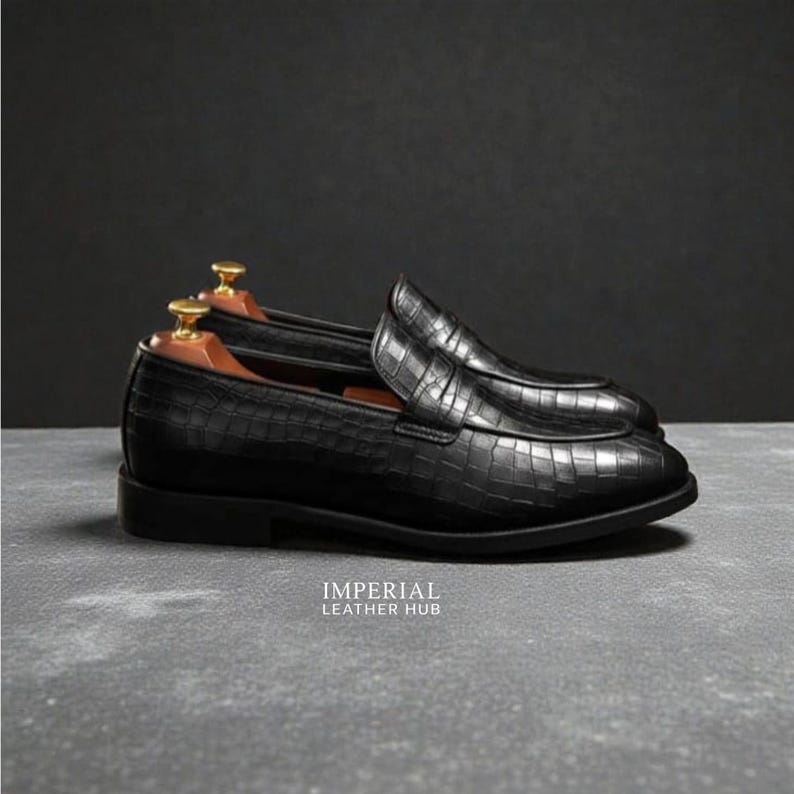 Pu&ograve; includere: Mocassini in pelle nera con effetto coccodrillo. Le scarpe hanno un design classico con tacco basso e punta arrotondata. Sono presenti forme per scarpe in legno. Il testo "IMPERIAL LEATHER HUB" &egrave; visibile.