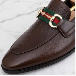 Könnte beinhalten: Nahaufnahme eines braunen Lederloafers mit goldfarbener Schnalle und einem grün-rot gestreiften Band. Der Schuh hat eine spitze Zehenpartie und eine strukturierte Lederoberfläche. Die Sohle ist schwarz.