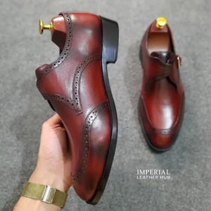 May include: A pair of deep burgundy leather monk strap shoes featuring brogue detailing along the seams, a dark stacked heel, and a gold-toned buckle. The shoes are displayed with wooden shoe trees inserted, and the text IMPERIAL LEATHER HUB is visible.