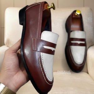 May include: A pair of two-tone loafers featuring a dark brown leather body with a white leather vamp and saddle strap. The shoes have white contrast stitching, a black sole, and a gold-toned shoe tree insert. The text IMPERIAL LEATHER HUB is visible.