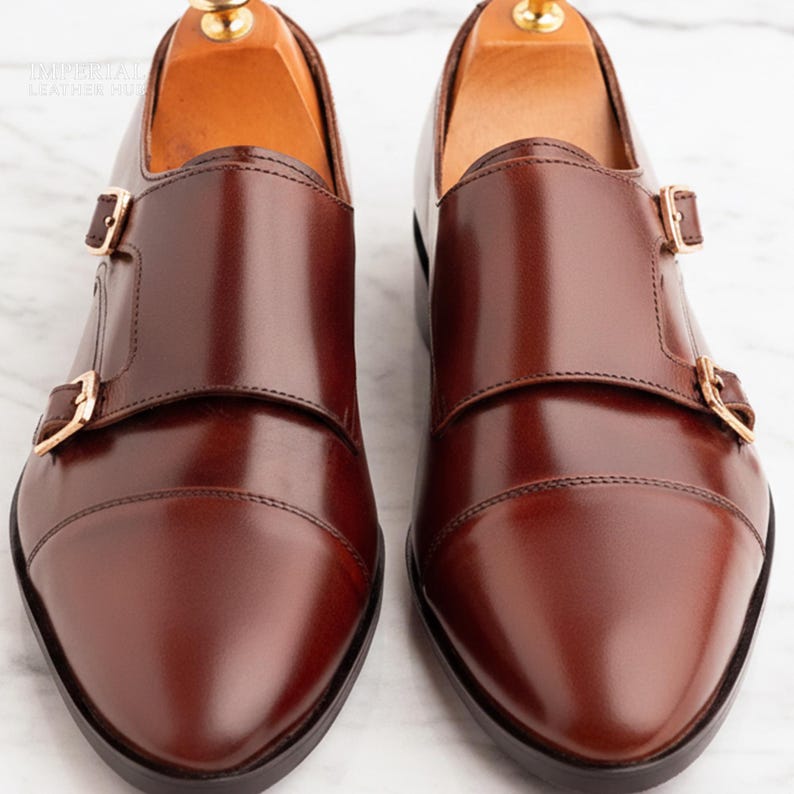 Peut inclure: Paire de chaussures &agrave; double boucle en cuir marron poli. Les chaussures ont un bout pointu et deux boucles dor&eacute;es. Elles sont pr&eacute;sent&eacute;es sur une surface en marbre blanc. Chaussures neuves et non port&eacute;es.
