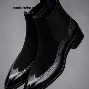 Op de afbeelding: Zwarte Chelsea-laarzen met een combinatie van glad leer en suède texturen. De laarzen zijn voorzien van een wingtip-neus met decoratieve brogue-perforaties en een treklipje bij de hiel. De tekst Imperial leather hut is zichtbaar.