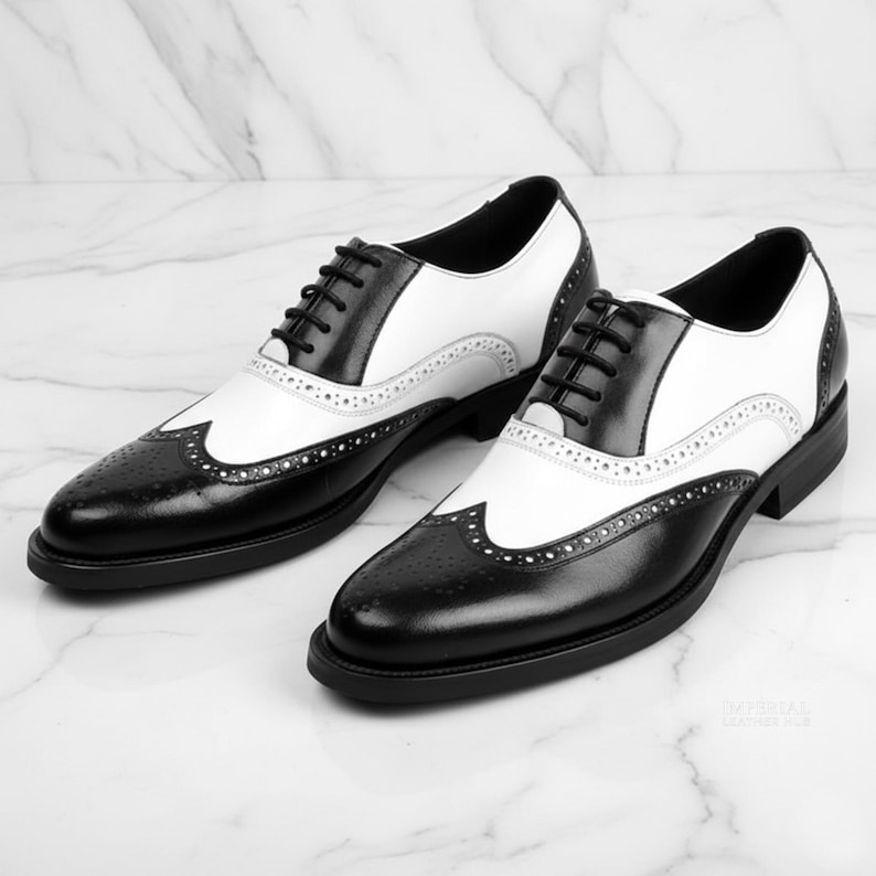 K&ouml;nnte beinhalten: Ein Paar zweifarbige Oxford-Schuhe aus schwarzem und wei&szlig;em Leder. Die Schuhe haben ein klassisches Fl&uuml;gelspitzendesign mit dekorativen Perforationen und schwarzen Schn&uuml;rsenkeln. Die Sohlen sind schwarz.