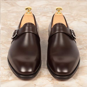 Könnte beinhalten: Ein Paar braune Leder-Monk-Schuhe mit silbernen Schnallen. Die Schuhe haben eine polierte Oberfläche und werden auf einer Marmoroberfläche präsentiert. Die Schuhe sind ein klassischer Stil, geeignet für formelle Anlässe.