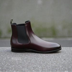 Pu&ograve; includere: Stivaletto Chelsea in pelle bordeaux lucida. Presenta un pannello laterale elastico nero e un tirante sul retro. La suola &egrave; nera. Il testo "IMPERIAL LEATHER HUB" &egrave; in basso. Design classico.