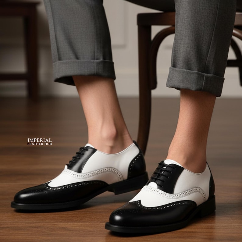 K&ouml;nnte beinhalten: Schwarz-wei&szlig;e Leder-Wingtip-Schuhe mit klassischem Design. Die Schuhe haben ein Lochmuster an den Kanten und einen Schn&uuml;rverschluss. Die Sohlen sind schwarz. Die Schuhe werden mit grauer Hose getragen.