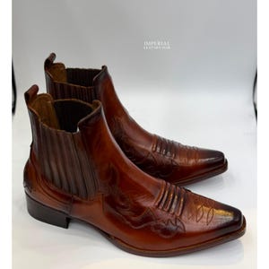 May include: A pair of brown leather Chelsea boots featuring a pointed toe, elastic side panels, and decorative stitching on the upper. The boots have a burnished finish and a pull tab at the heel. The text IMPERIAL LEATHER HUB is visible.