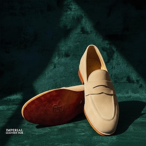 May include: Beige suede penny loafers with a brown leather sole. The shoes are displayed on a dark green surface. The sole of one shoe is visible, showing a logo. The words "IMPERIAL LEATHER HUB" are at the bottom left.