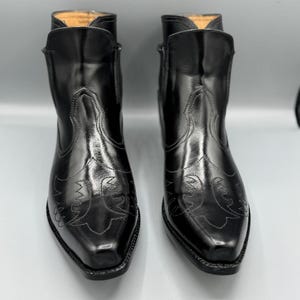 Puede incluir: Un par de botines de cuero negro con punta afilada, tacón bajo de bloque y costuras decorativas en tono sobre tono en el empeine. Los botines tienen un acabado liso y pulido, con una caña estructurada y un diseño de borde superior.