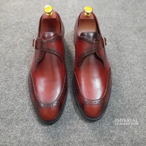 May include: A pair of dark brown leather monk strap shoes featuring brogue detailing along the toe cap and strap, a single gold-tone buckle, and a gold-tone shoe tree insert. The text IMPERIAL LEATHER HUB is visible in the bottom right corner.