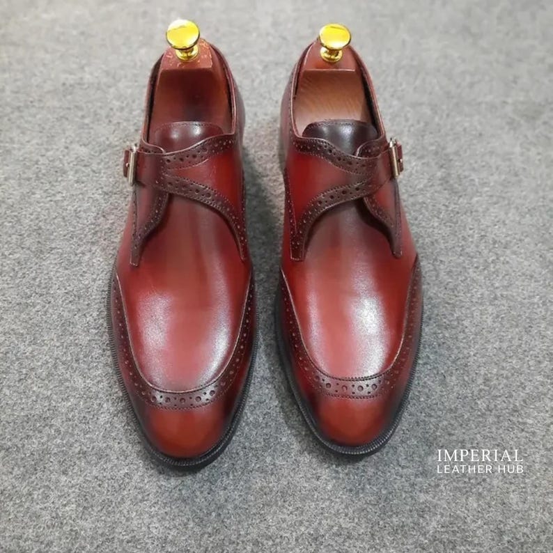 May include: A pair of dark brown leather monk strap shoes featuring brogue detailing along the toe cap and strap, a single gold-tone buckle, and a gold-tone shoe tree insert. The text IMPERIAL LEATHER HUB is visible in the bottom right corner.