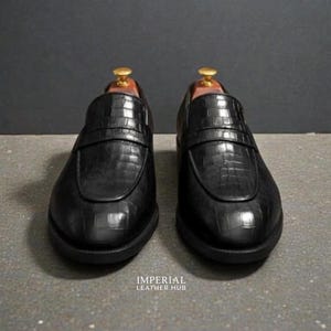 Pu&ograve; includere: Un paio di mocassini neri in pelle con una texture a pelle di coccodrillo. Le scarpe hanno un design classico slip-on con una cinghia sulla parte superiore. Le suole sono nere e le scarpe sono esposte su una superficie grigia. Il testo "IMPERIAL LEATHER HUB" &egrave; visibile.