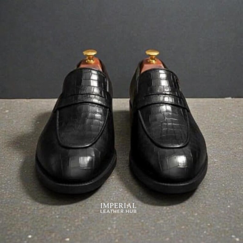 Pu&ograve; includere: Un paio di mocassini neri in pelle con una texture a pelle di coccodrillo. Le scarpe hanno un design classico slip-on con una cinghia sulla parte superiore. Le suole sono nere e le scarpe sono esposte su una superficie grigia. Il testo "IMPERIAL LEATHER HUB" &egrave; visibile.
