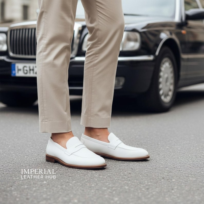 K&ouml;nnte beinhalten: Wei&szlig;e Leder-Loafer mit braunen Sohlen. Die Schuhe werden mit beigefarbenen Hosen getragen. Das Bild wird im Freien mit einem schwarzen Auto im Hintergrund aufgenommen. Der Text "IMPERIAL LEATHER HUB" befindet sich unten.