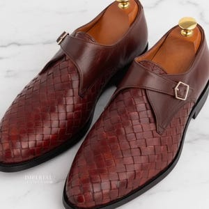 May include: A pair of brown leather monk strap shoes with a woven design. The shoes feature a buckle fastening and are presented on a white marble surface. The shoe's interior is a lighter shade of brown.