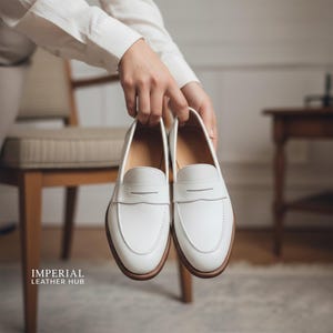 K&ouml;nnte beinhalten: Ein Paar wei&szlig;e Leder-Loafer mit klassischem Design. Die Schuhe haben eine runde Spitze, einen Riemen &uuml;ber dem Spann und eine braune Sohle. Die Schuhe werden von einer Person gehalten. Der Text "IMPERIAL LEATHER HUB" befindet sich in der unteren linken Ecke.