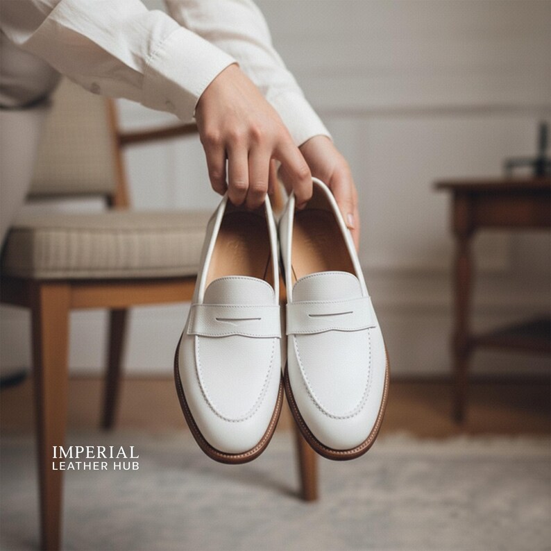 K&ouml;nnte beinhalten: Ein Paar wei&szlig;e Leder-Loafer mit klassischem Design. Die Schuhe haben eine runde Spitze, einen Riemen &uuml;ber dem Spann und eine braune Sohle. Die Schuhe werden von einer Person gehalten. Der Text "IMPERIAL LEATHER HUB" befindet sich in der unteren linken Ecke.