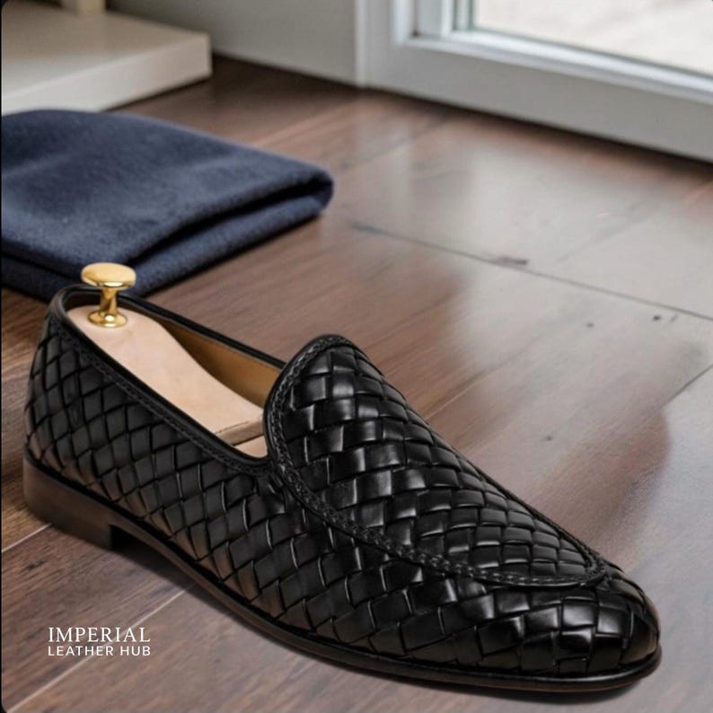 May include: Black woven leather loafer with a wooden shoe tree inside. The shoe is on a wooden floor, with a folded blue cloth in the background. The shoe has a rounded toe and a low heel. The text "IMPERIAL LEATHER HUB" is visible.