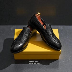 Pu&ograve; includere: Un paio di mocassini in pelle nera con una texture a pelle di coccodrillo, appoggiati su una scatola gialla. Le scarpe hanno un classico design slip-on. Il testo "IMPERIAL LEATHER HUB" &egrave; visibile sulla scatola.