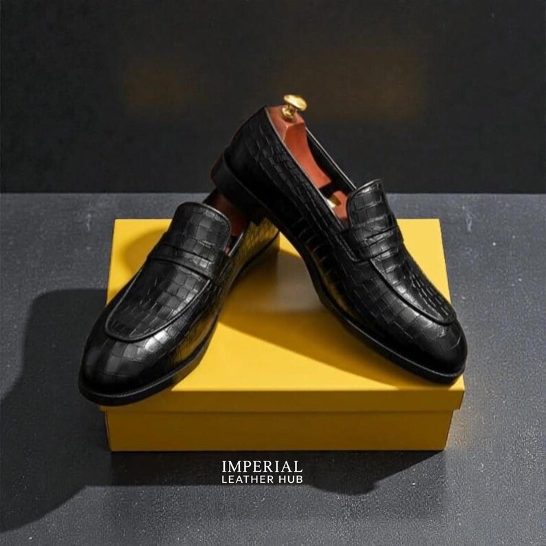 Pu&ograve; includere: Un paio di mocassini in pelle nera con una texture a pelle di coccodrillo, appoggiati su una scatola gialla. Le scarpe hanno un classico design slip-on. Il testo "IMPERIAL LEATHER HUB" &egrave; visibile sulla scatola.