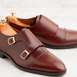 May include: A pair of brown leather double monk strap shoes with gold-coloured buckles. The shoes are displayed on a white marble surface. The shoes have a polished finish and are designed for formal wear.