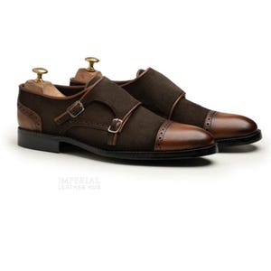 May include: A pair of brown leather double monk strap shoes. The shoes feature a dark brown suede upper with leather accents, including the toe cap and straps. The shoes have a classic design with a buckle closure and are displayed on wooden shoe trees.
