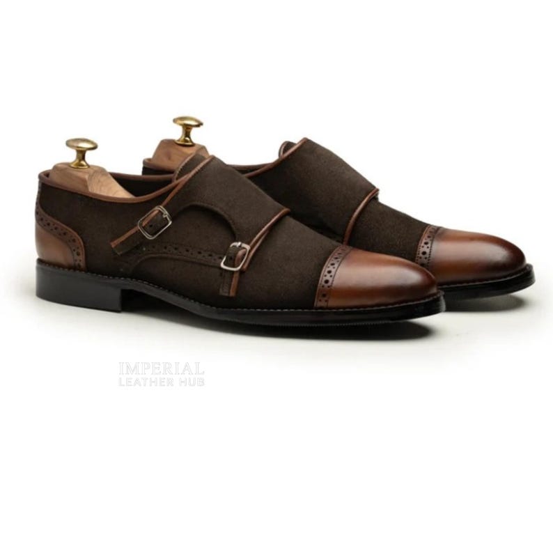 May include: A pair of brown leather double monk strap shoes. The shoes feature a dark brown suede upper with leather accents, including the toe cap and straps. The shoes have a classic design with a buckle closure and are displayed on wooden shoe trees.
