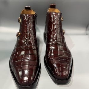 May include: Burgundy leather ankle boots with a textured, checkered pattern. The boots have a pointed toe, a side zip, and two gold-coloured buckles. The boots are displayed on a white surface.