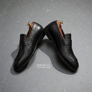 Pu&ograve; includere: Un paio di mocassini in pelle nera con una texture a coccodrillo. Le scarpe hanno un design classico con un cinturino sulla parte superiore e sono esposte con forme di legno. Il testo "IMPERIAL LEATHER HUB" &egrave; visibile.