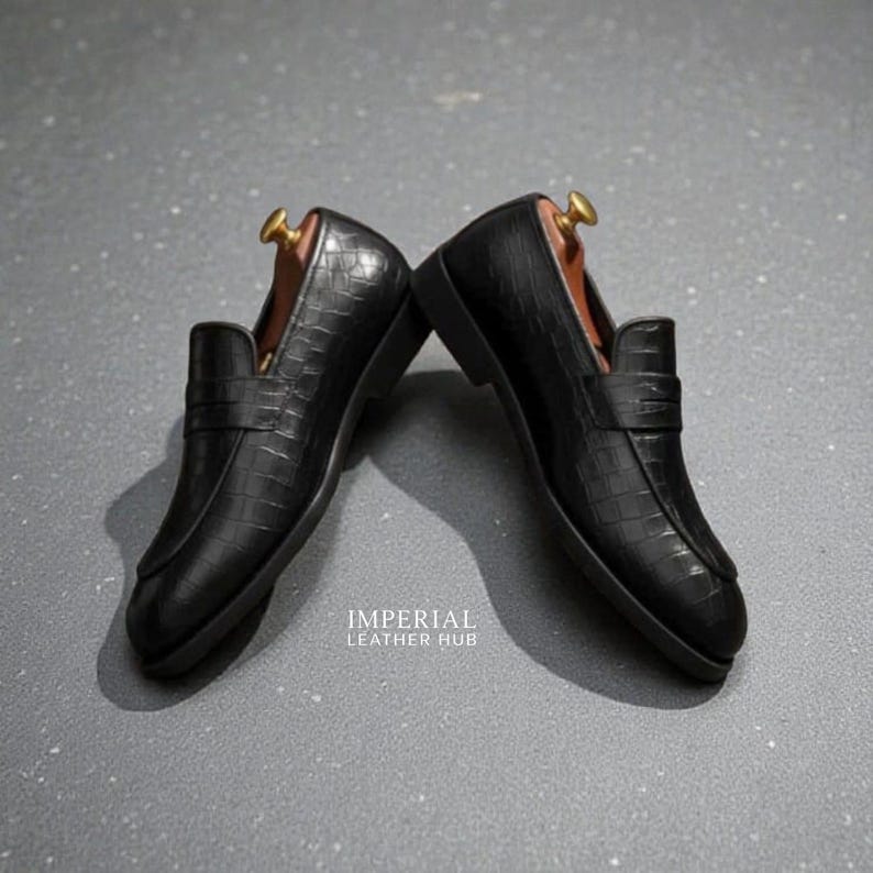 Pu&ograve; includere: Un paio di mocassini in pelle nera con una texture a coccodrillo. Le scarpe hanno un design classico con un cinturino sulla parte superiore e sono esposte con forme di legno. Il testo "IMPERIAL LEATHER HUB" &egrave; visibile.