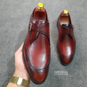 May include: A pair of deep burgundy leather monk strap shoes featuring a single buckle closure, intricate brogue detailing along the toe cap and strap, and a wooden shoe tree insert. The text IMPERIAL LEATHER HUB is visible in the lower right corner.
