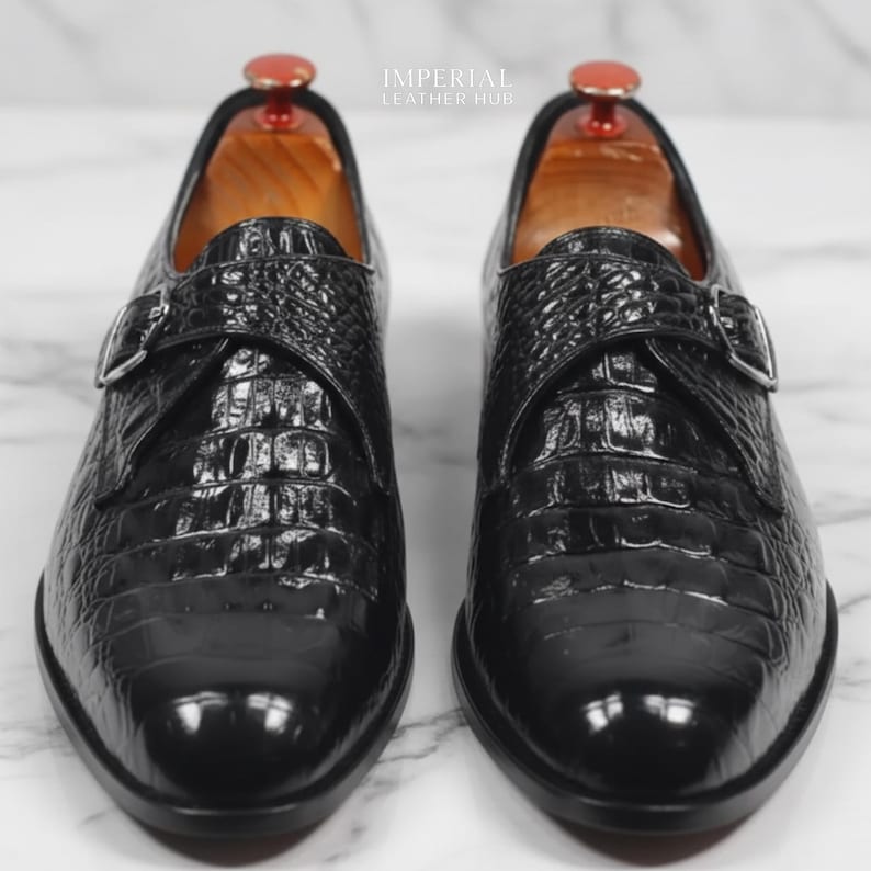 May include: A pair of black leather dress shoes with a crocodile skin texture. The shoes have a buckle closure and are displayed on wooden shoe trees. The image includes the text "IMPERIAL LEATHER HUB".