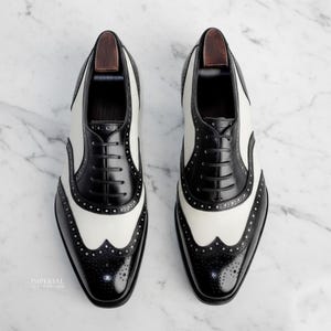 K&ouml;nnte beinhalten: Zweifarbige Wingtip-Schuhe in Schwarz und Wei&szlig; mit klassischem Brogue-Design. Die Lederschuhe haben einen Schn&uuml;rverschluss. Das Bild zeigt die Schuhe auf einer Marmoroberfl&auml;che. Stilvolle Herrenschuhe.
