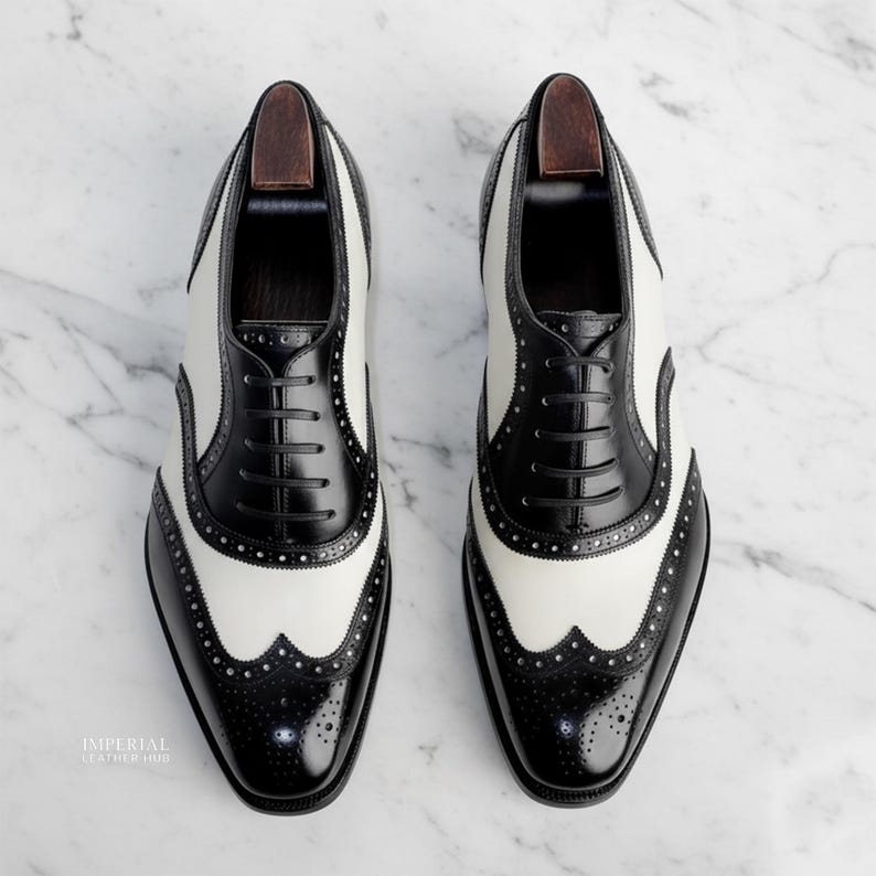 K&ouml;nnte beinhalten: Zweifarbige Wingtip-Schuhe in Schwarz und Wei&szlig; mit klassischem Brogue-Design. Die Lederschuhe haben einen Schn&uuml;rverschluss. Das Bild zeigt die Schuhe auf einer Marmoroberfl&auml;che. Stilvolle Herrenschuhe.