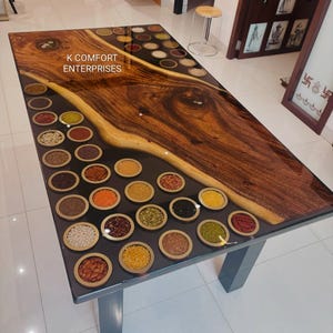 Può includere: Un tavolo rettangolare in legno con base grigio scuro. La superficie del tavolo presenta un design a venature naturali con finitura lucida. Piccole ciotole di legno riempite con vari cereali e semi sono incorporate. Il testo "K COMFORT ENTERPRISES" è visibile.