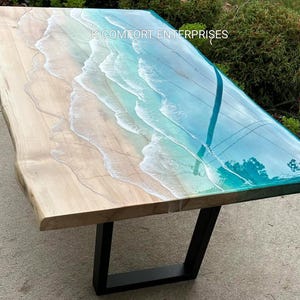 Op de afbeelding: Een houten tafel met een epoxyharsontwerp met strandthema. De tafel heeft een licht houten oppervlak dat overgaat in een blauw en wit harsontwerp, dat oceaangolven nabootst. De tafel wordt ondersteund door zwarte metalen poten en de tekst "K COMFORT ENTERPRISES" is zichtbaar.