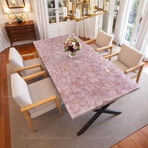 Op de afbeelding: Een rechthoekige eettafel met een roze, patroon oppervlak, ondersteund door een zwarte metalen basis. De tafel is omringd door vier beige gestoffeerde stoelen met houten armleuningen. Een vaas met bloemen staat op de tafel.