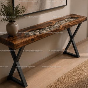 Puede incluir: Una consola de madera con acabado oscuro y patas de metal negro. La encimera presenta una incrustación de resina transparente llena de guijarros grises lisos. Un jarrón con vegetación se encuentra en el lado izquierdo.
