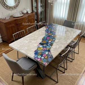 May include: A rectangular dining table with a white surface and a central river of colourful agate stones. The table has dark metal legs and is surrounded by grey chairs. A chandelier hangs above.
