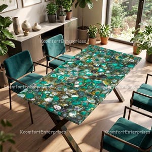 May include: A rectangular dining table with a green and white agate stone top. The table is supported by dark brown, crisscross legs. Four teal velvet chairs with dark metal frames surround the table. The room has large windows and potted plants.