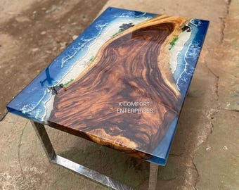 May include: A rectangular coffee table with a wooden top and metal legs. The table's surface features a unique design with a wood grain center, surrounded by a blue resin border that mimics a beach scene. The table is supported by silver metal legs.