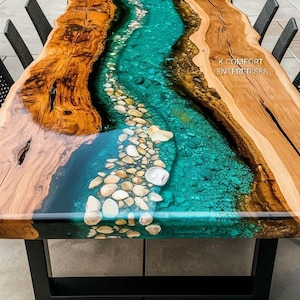 Op de afbeelding: Een handgemaakte houten tafel met een rivierontwerp van epoxyhars. De tafel heeft een turquoise hars middenstuk met ingebedde stenen, omlijst door natuurlijke houten randen. Hij staat op een zwart metalen frame. De tekst "K COMFORT ENTERPRISES" is zichtbaar.