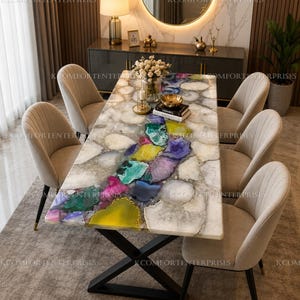 May include: A rectangular dining table with a white stone surface featuring a central inlay of multicoloured agate slices in shades of purple, yellow, green, and blue. The table has a black metal X-shaped base and is surrounded by six beige chairs.