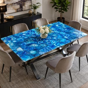 Op de afbeelding: Een rechthoekige eettafel met een levendig blauw agaatstenen blad. De tafel wordt ondersteund door een zwart metalen onderstel en is omringd door zes grijze gestoffeerde stoelen. Een vaas met witte bloemen staat op de tafel.
