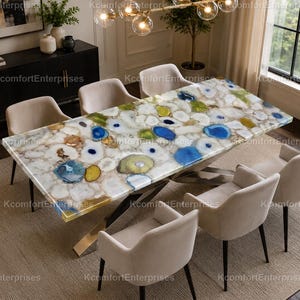 May include: A rectangular dining table with a white and multi-coloured agate stone top. The table has a gold-coloured metal base and is surrounded by six beige upholstered chairs. The room has a neutral colour palette.