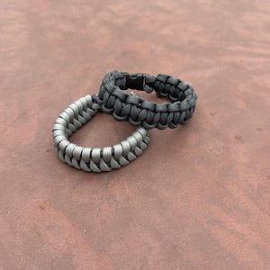 May include: Two paracord bracelets. One is a light gray color, and the other is a darker gray. Both bracelets are made of woven cord and have a plastic buckle closure. The bracelets are shown on a brown surface.